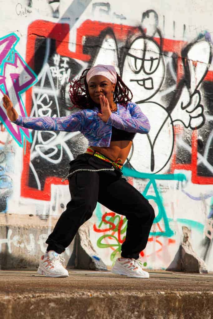 A black-african cis woman dancing in front of colourful graffiti on a wall. She is and wearing a light pink head scarf over braided hair with a purple top, black trousers and white trainers. 