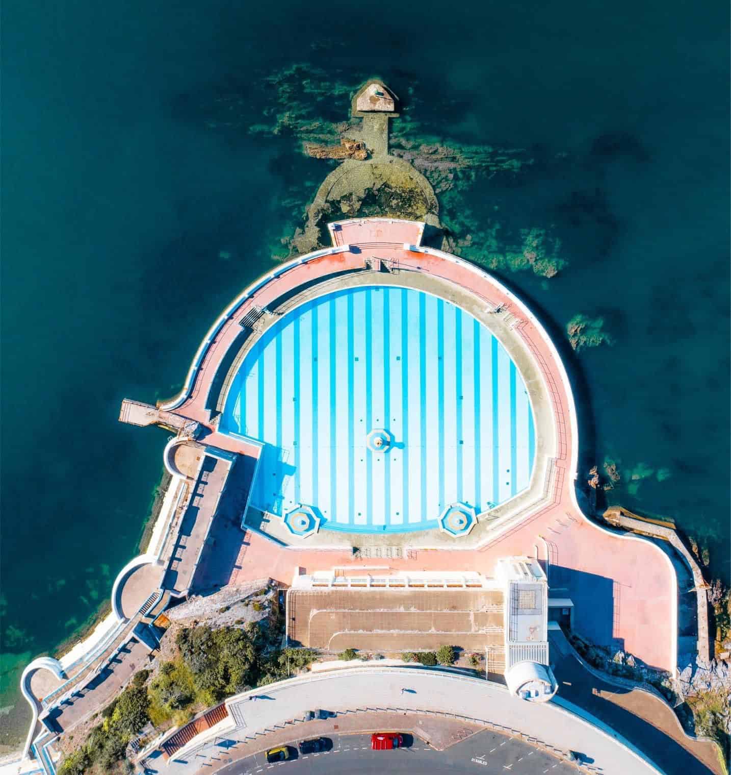 An aerial photo of a round outdoor lido swimming pool jutting out into the sea - the pool has light blue stripes on the bottom and light pink paths around it. The pool is in the art deco style and has three fountains. The sea around the pool is a dark blue colour.